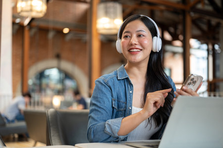 Happy asian woman wearing headphones looking away from phone and laptop sitting at table in the cafe. Listening to Music, Leisure Time, Urban City Life.の写真素材