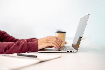 A man student's hands typing on laptop aside notebook and coffee on white table in a cafe or...
