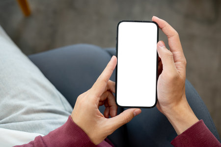 A person's hands holding white screen smartphone with both hands while sitting indoors at a cafe or library. Modern Technology, Urban Life, Advertising Mockup.の写真素材
