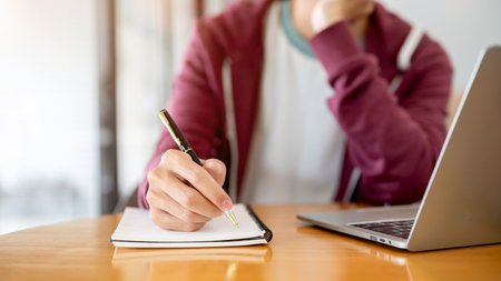 A man student's hand holding pen writing in notebook aside laptop on wooden table in cafe or...