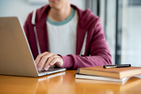Close up of a man student typing on laptop aside books and pen on wooden table in a cafe or library. Working or Study, Urban Life, Modern Technology.の写真素材