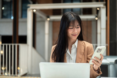 Asian businesswoman or office worker holding or looking at smartphone over laptop on table in a cafe. Professional Suit, Remote Working, Daily Life.の写真素材