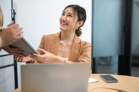 Asian businesswoman receive tablet from coworker as sitting at wooden table in office meeting room....
