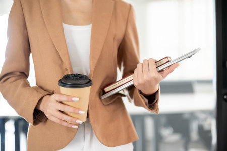 Close up businesswoman office worker holding coffee with book on laptop standing walking in building. Working Hours, Corporate Desk Job, Modern Company.の写真素材