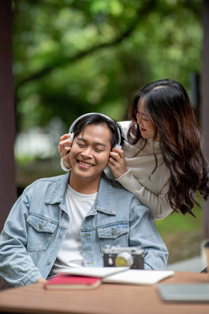 Young asian couple a standing woman putting headphones on man while sitting at outdoor table in park. Dating activity, Urban Life, Chilling Outdoorsの写真素材