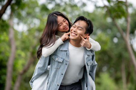 Young asian couple fooling around woman poking man's cheeks while getting piggyback walking in park....