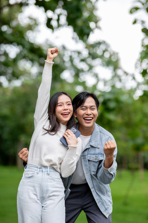 Young asian couple making a fist with woman holding phone standing outdoors in park. Dating activity, Nature Healing, Chilling Outside.の写真素材