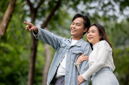 Young asian couple cuddling woman hugging arm of pointing man standing or walking outdoors in park. Dating activity, Nature Healing, Chilling Outside.の写真素材