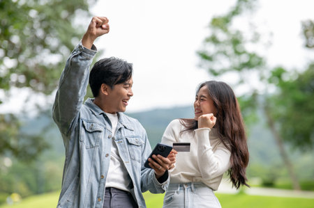 Young asian couple man with phone raising fist a woman holding card standing outdoor in park.の写真素材