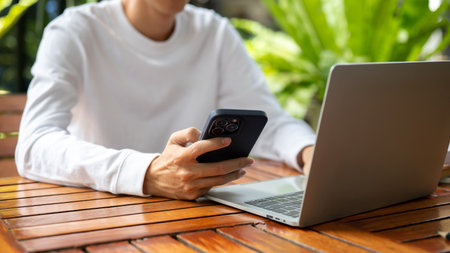 Close up of a man's hand holding phone aside laptop while sitting outdoors at wooden table in cafe. Working or Study, Urban Life, Modern Technology.の写真素材