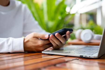 Close up of man's hands holding using phone over laptop while sitting outdoor at wooden cafe table. Working or Study, Urban Life, Modern Technology.の写真素材