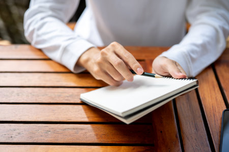 Close up of a man's hand holding pencil sketching in notebook while sitting outdoors at wooden...