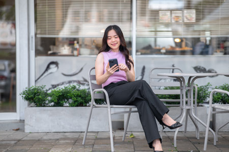 Young asian woman holding and looking at phone with smile while sitting at table in front of a cafe. Modern Tech, Urban Life, Chilling Outdoors.の写真素材