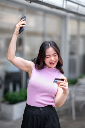 Young asian woman looking at credit card raising a hand with phone while standing in front of a cafe. Bank advertising, Urban Life, Chilling Outdoors.の写真素材