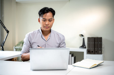 Asian businessman office worker sitting holding pen looking at laptop aside notebook on white table. Working Hours.の写真素材