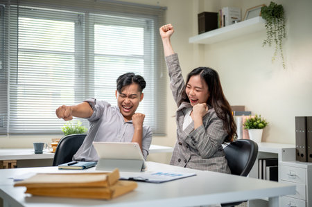Excited businessman and woman coworker raising hand while looking at tablet on table in office.