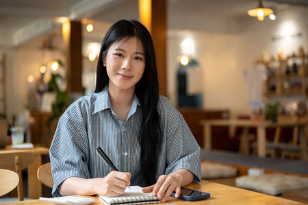 Asian woman smiling while holding pen over a notebook with calculator and phone aside on wooden...