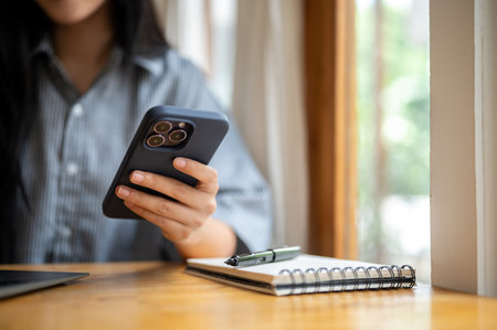 Close up of woman's hand holding or touching on smartphone with pen atop notebook and laptop at cafe...