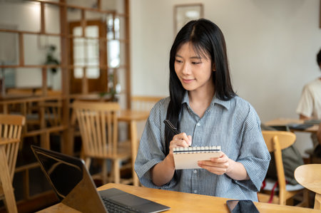 Smiling asian woman holding notebook and writing with pen while looking at laptop on wooden table in the cafe. Working or Study. Chilling Outside.の写真素材