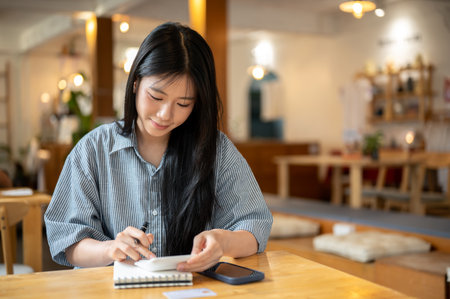 Asian woman holding pen and using calculator over notebook with calculator and phone aside on cafe...