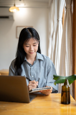 Smiling woman holding pen writing in notebook with laptop and glass bottle vase aside on cafe's...