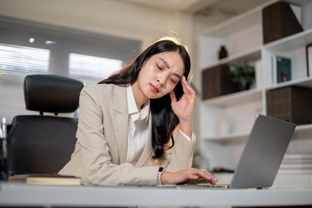 Asian businesswoman or female office worker massaging temple getting headache while typing on laptop...