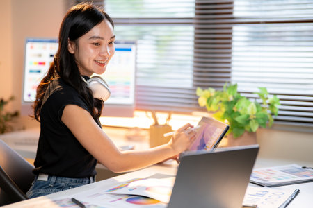 Asian woman designer drawing with stylus on tablet looking at laptop as sitting with color chart at...
