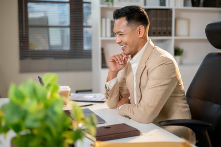 Smiling asian businessman holding his chin and looking happily at laptop with notebook aside on office's working table. Boss or Manager, Company jobs.の写真素材