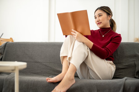 Pretty asian woman holding and reading a book while sitting knee up on sofa in a living room or...