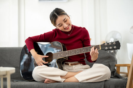 Pretty asian woman holding and playing a guitar while sitting cross legged on sofa in a living room...