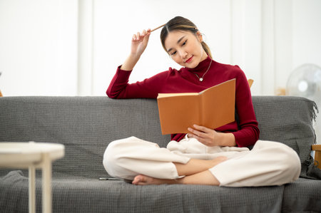 Pretty asian woman holding pencil on head and reading a book while sitting on sofa in a living room...