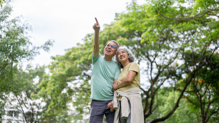 Old woman standing hugging glasses old man's arm while he's pointing away on grass lawn in park....