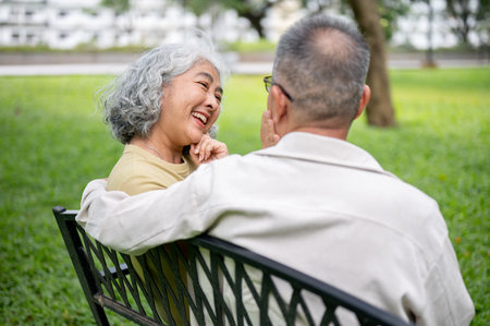 Close up of an old woman smiling talking to a glasses old man while sitting together on bench in park. Elderly couple, Nature park, Chilling Outdoor.の写真素材