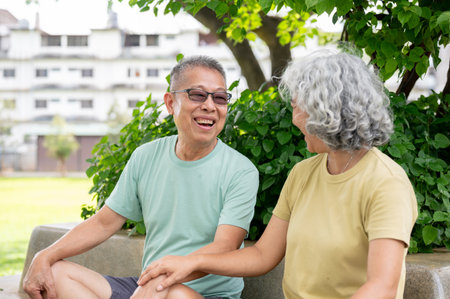 Glass old man smiling and talking with old woman while sitting together on cement bench in the park....