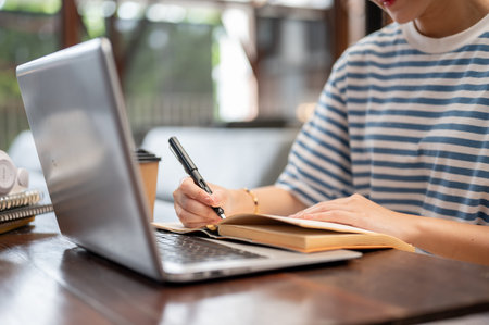 Close up of a woman writing in notebook with a fountain pen while sits across laptop on table in...