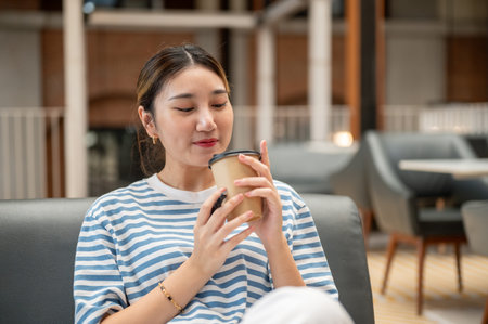 Asian young woman holding and smelling coffee aroma inside a cup while sitting on sofa in the cafe. Leisure free time.の写真素材