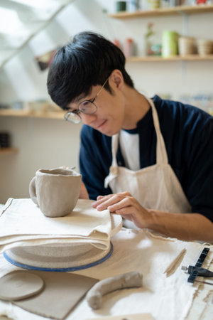 Young man potter artist adding texture on a clay mug at working table in a pottery class or studio....