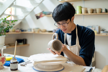 Young man potter artist holding and looking at clay mug at working table in a pottery class or...