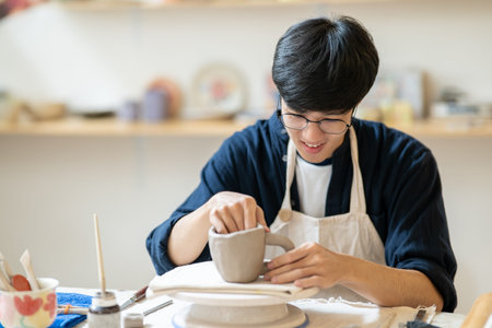 Young man pottery artist molding clay mug cup on throwing wheel at working table in a class or...