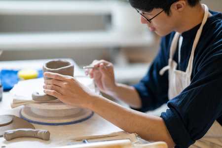 Potter artist holding brush adding texture on a clay mug on working table in a pottery class or...