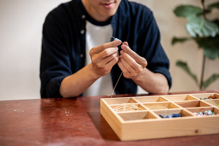 Young man artist threading ceramic beads making a colorful bracelet aside wooden box in a class or...