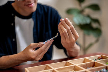 Young man artist holding and looking at ceramic beads on thread while making a bracelet in a class...