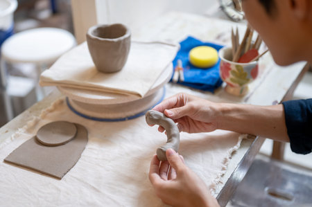Close up of pottery artist's hands molding clay into a mug handle at working table in class or...