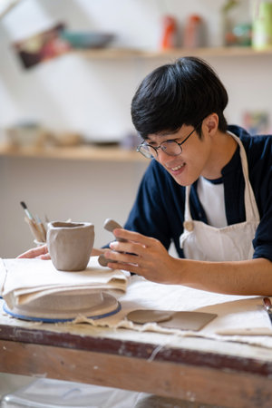 Potter artist holding and adding handle to clay mug cup on working table in a pottery class or...