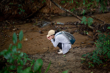 Asian woman tourist crouching aside water stream to drink or wash her face during forest hiking or...
