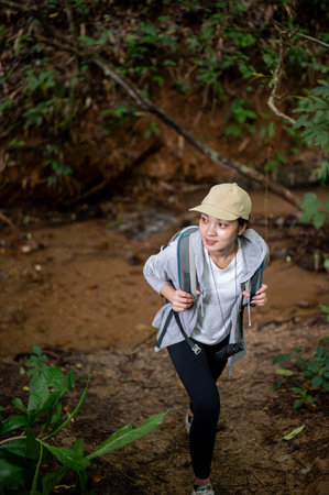 Asian woman tourist holding bag straps and walking up from a water stream during forest hiking or...