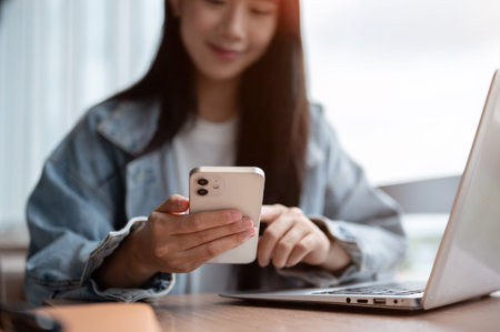 Close up of woman in denim jacket holding and looking at smartphone over laptop on wooden table in...