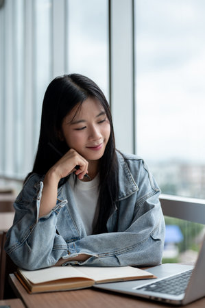 Young asian woman in denim jacket rest head on a hand and looking at laptop on wooden table in cafe....