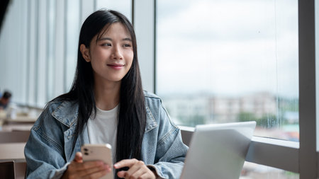 Young asian woman in denim jacket holding phone as she smiling look away aside laptop on table in...
