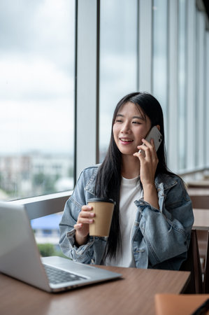 Young asian woman in denim jacket smiling and holding phone talking aside laptop on table in cafe....